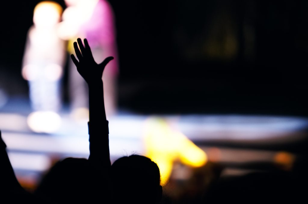 Supporter in the audience raising a hand at a live performance silhouetted against the blurred outline of people on stage
