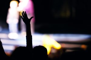 Supporter in the audience raising a hand at a live performance silhouetted against the blurred outline of people on stage