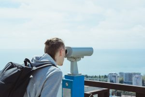 young hipster tourist man looking through metal coin operated binoculars on sea, sky and city, horizontal lifestyle stock photo image